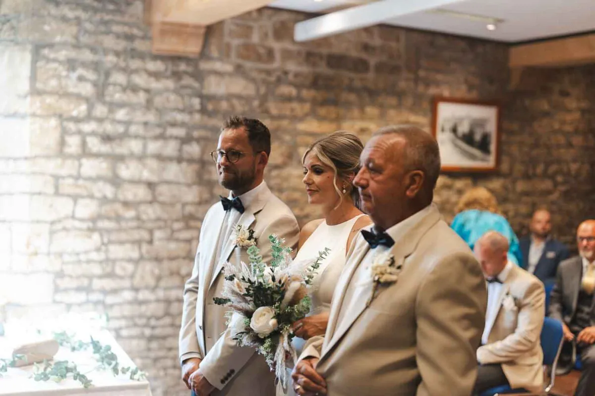 A bride standing beside her groom in a beige suit and her father during a civil ceremony inside a stone barn venue.
