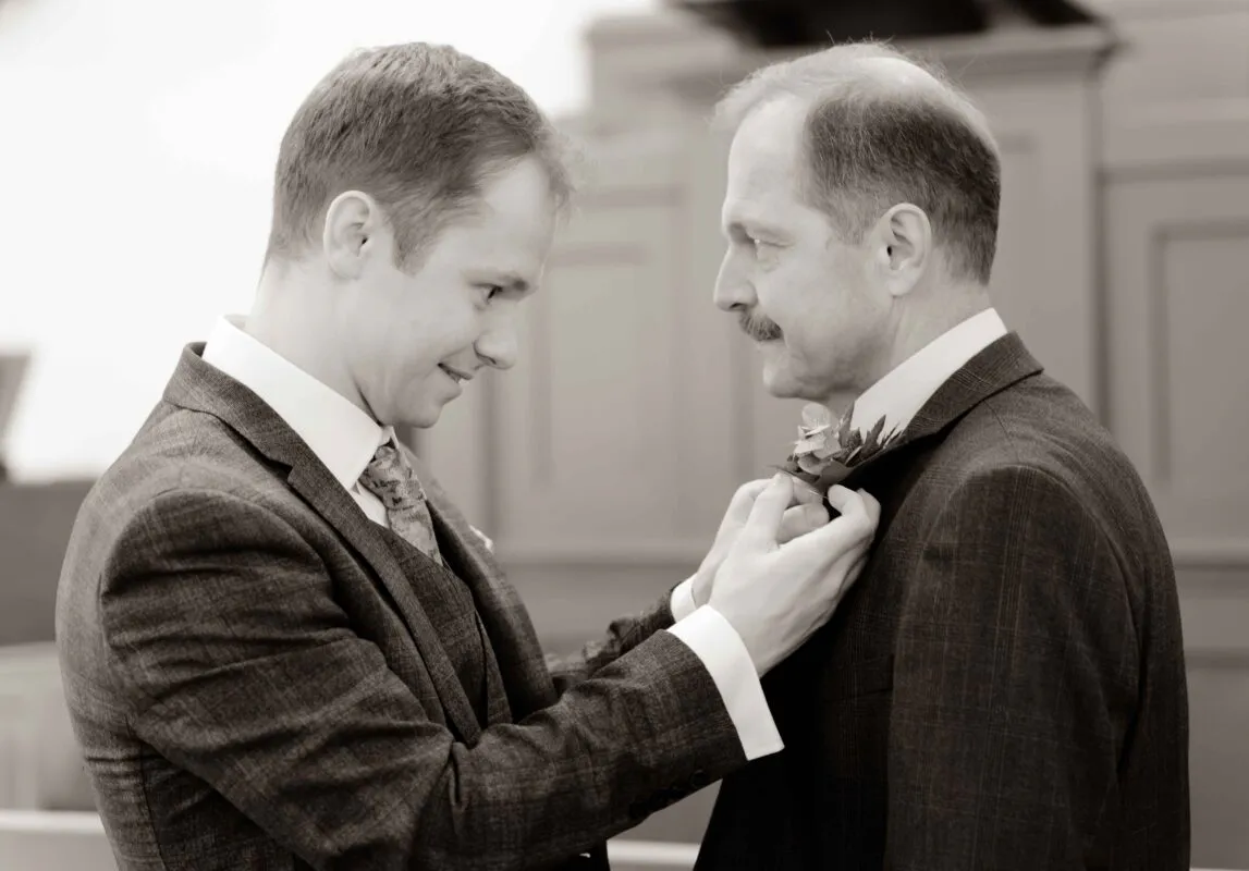 A groom in a patterned three-piece suit carefully attaches a boutonniere to the lapel of another man in a matching suit.