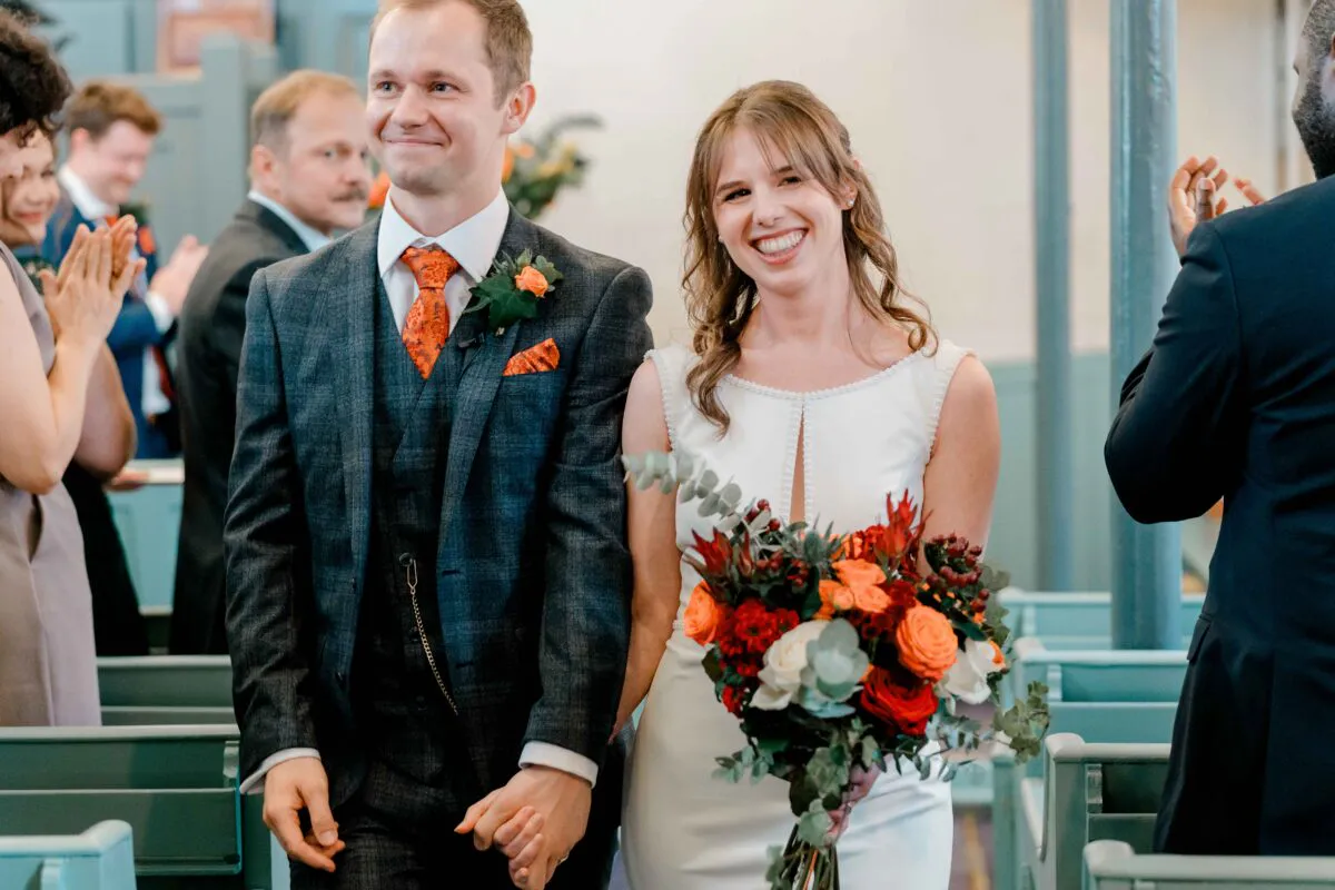 A smiling bride and groom walk hand in hand down the aisle of a church after their ceremony, holding a vibrant bouquet of orange and red flowers.
