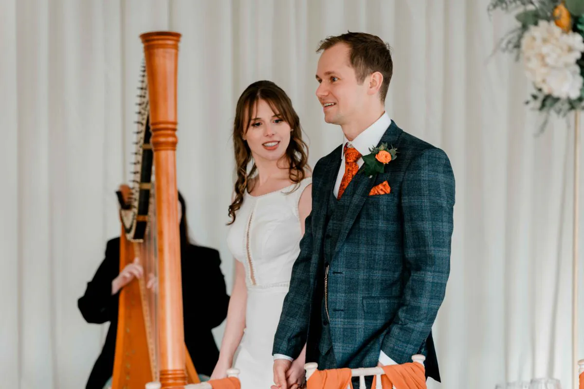 A smiling groom in a checked suit and a bride in a white dress stand together during a wedding ceremony, with a harpist playing in the background.