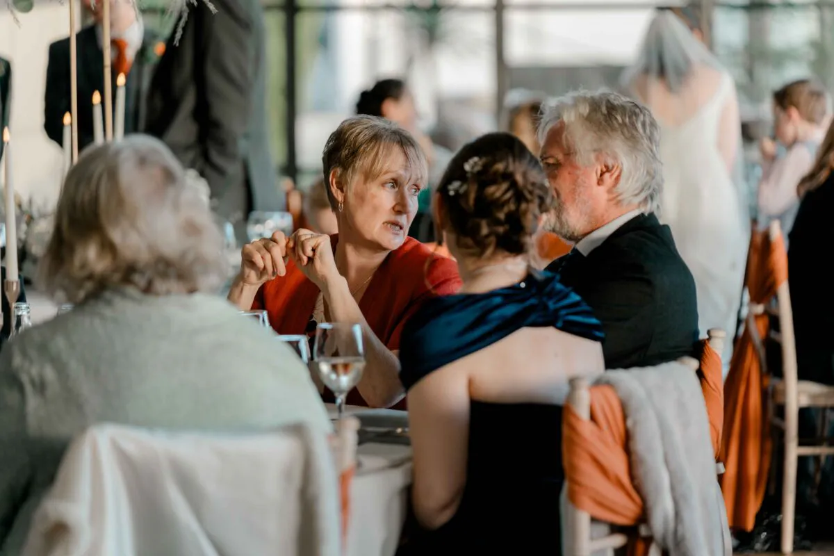 Wedding guests converse animatedly around a brightly lit dining table during the wedding reception.