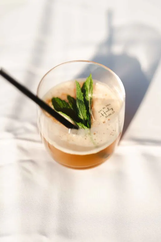 A refreshing signature wedding cocktail garnished with fresh mint leaves and served in a personalised glass on a sunlit table.