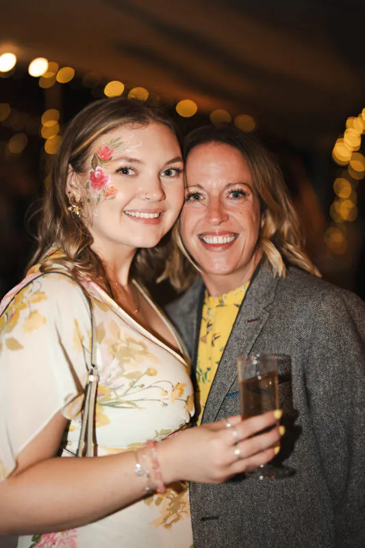 Two wedding guests smiling together at the evening reception, with one guest sporting vibrant floral face paint and holding a glass of champagne.