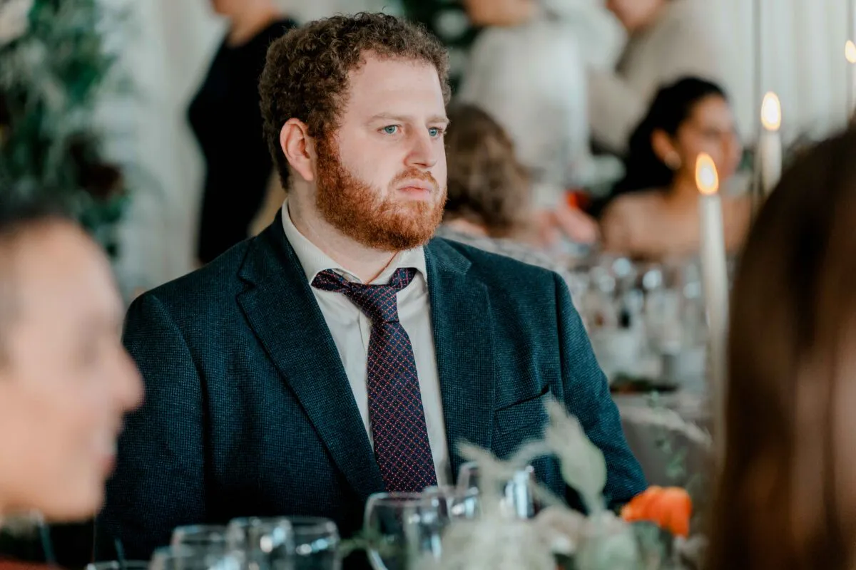 A wedding guest with curly brown hair and a beard sits attentively at a candlelit reception table during the speeches.