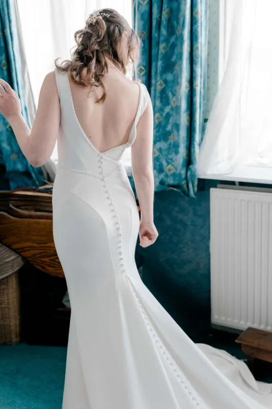 A bride stands from the back showcasing the elegant low scoop back and fabric covered button details of her fitted white wedding gown.