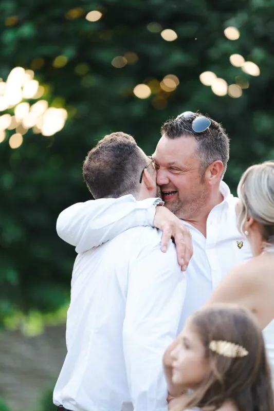 Two grooms in white shirts share a joyful embrace outdoors while laughing together against a backdrop of soft green foliage.
