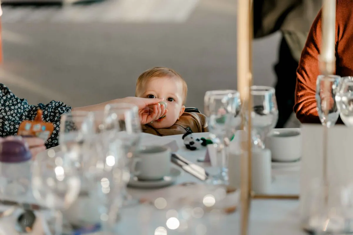 A young baby sits quietly at a wedding reception table surrounded by glassware and place settings during the wedding breakfast.
