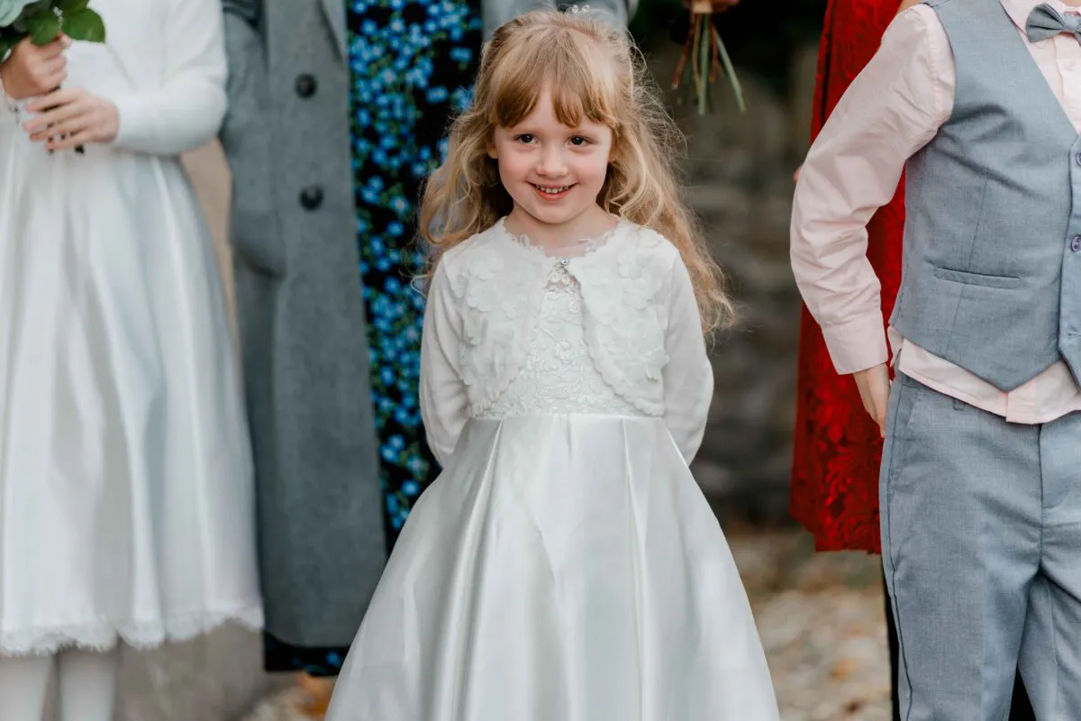 A young flower girl smiling warmly while wearing a white dress and matching cardigan during an outdoor wedding celebration.