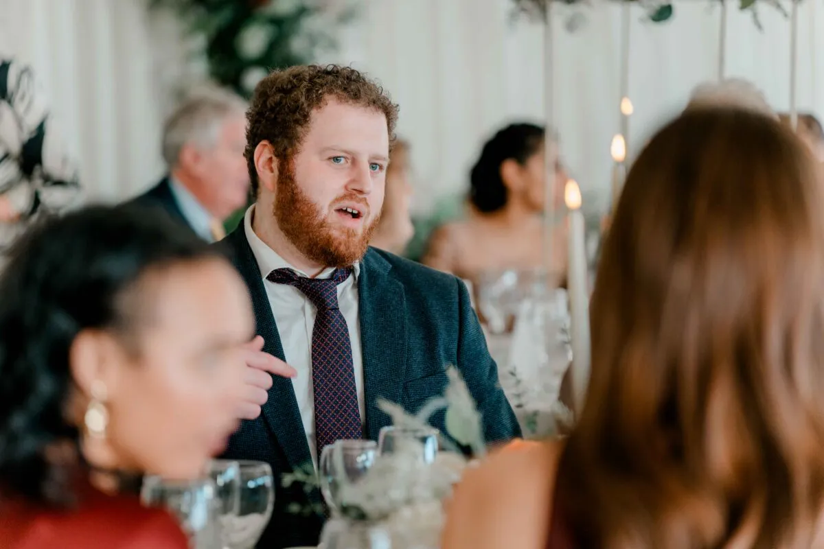 A candid photograph captures a bearded groom speaking animatedly during the wedding breakfast while seated at a candlelit table among guests.
