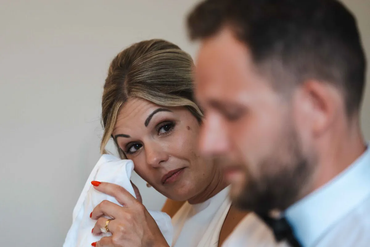 A tearful bride wipes away an emotional tear with a handkerchief while looking over at the groom during an intimate wedding moment.