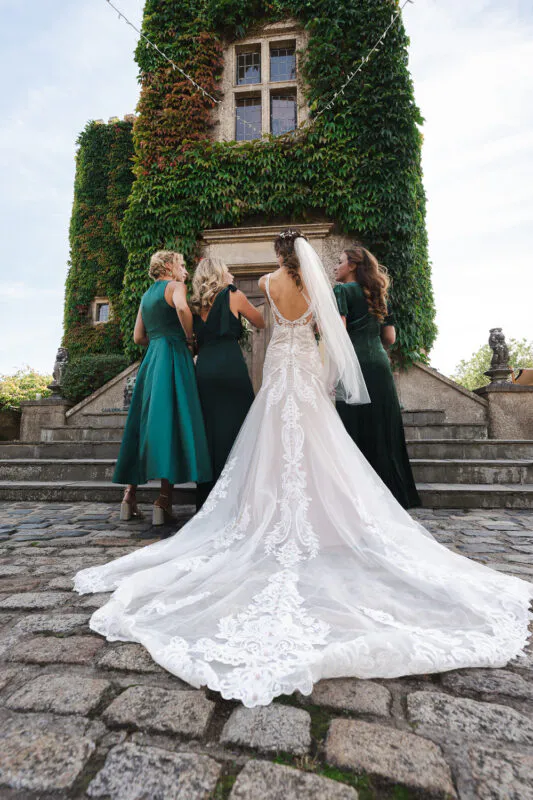 A bride in an intricate lace gown with a long train stands with her bridesmaids in emerald green dresses in front of an ivy-covered stone tower.