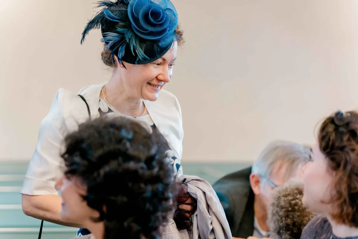 A smiling wedding guest wearing a blue fascinator and white jacket mingles with other guests during the wedding reception.