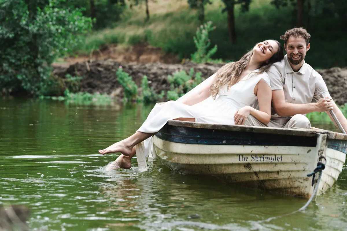 A bride and groom share a joyful, laughing moment together while relaxing in a small wooden boat on a calm lake.
