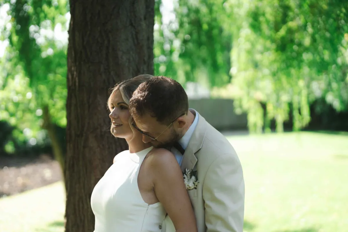 A groom gently kisses the bride's shoulder outdoors against a lush green woodland backdrop with dappled sunlight.