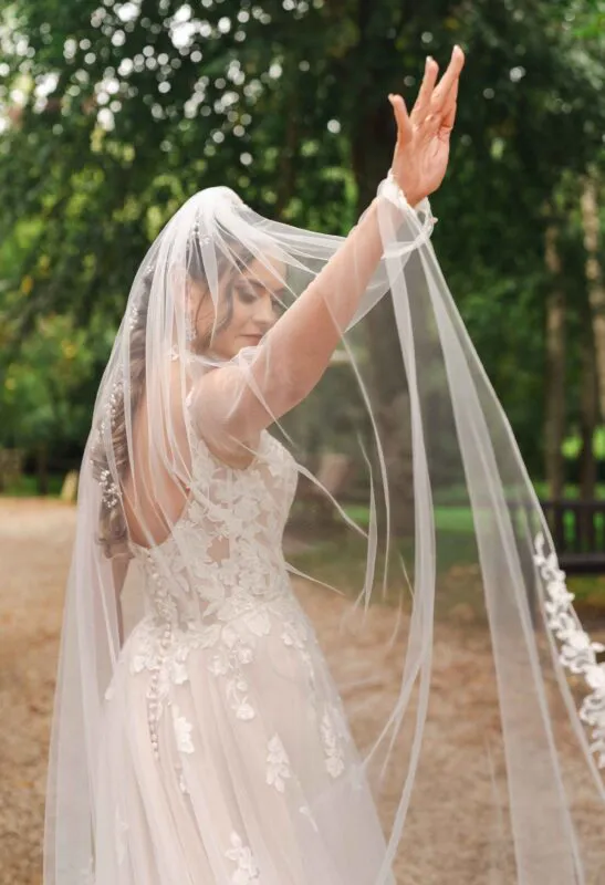 A bride in a lace gown raises her arm, creating graceful movement with her long veil against a lush woodland backdrop.