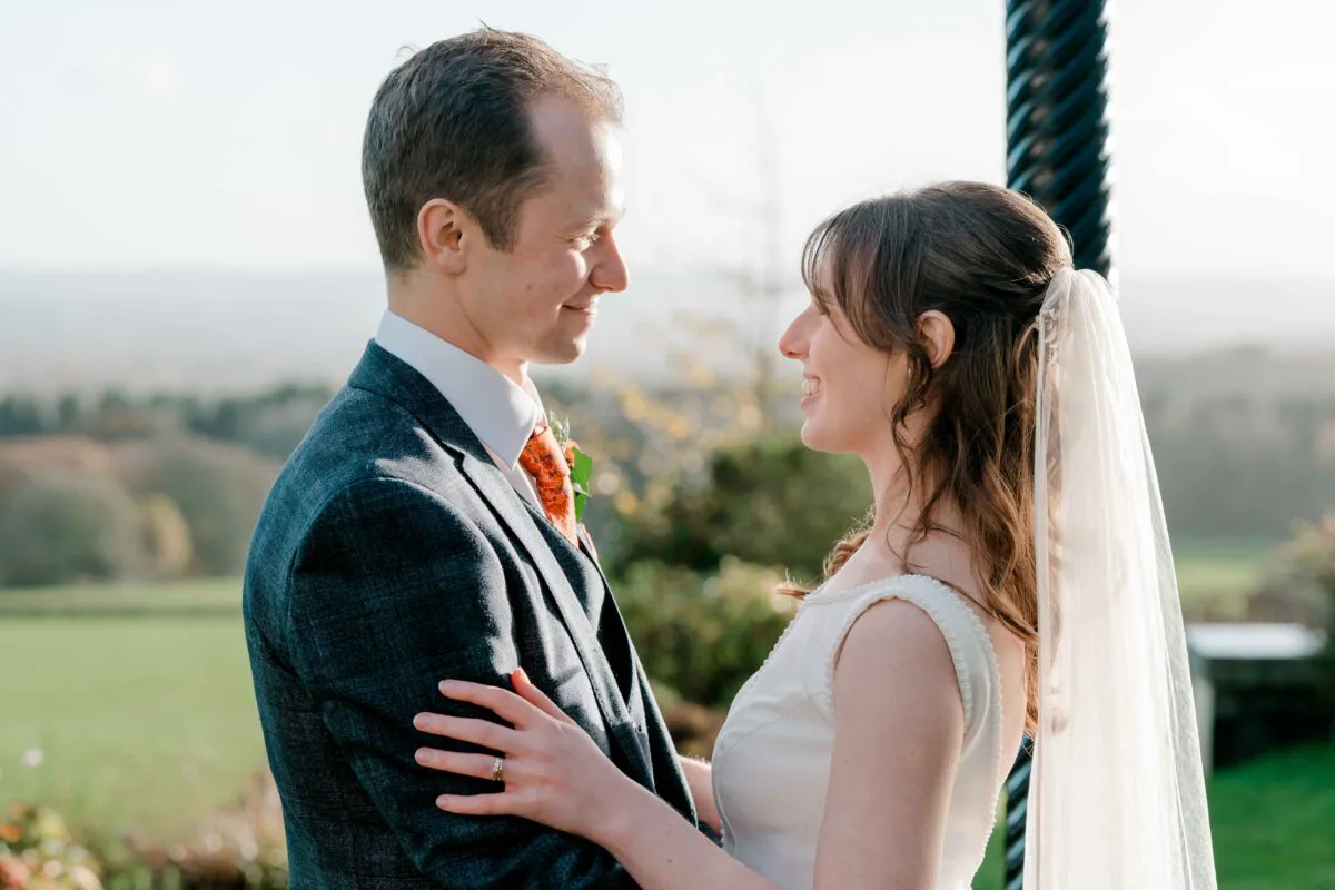 A smiling bride and groom gaze lovingly at each other outdoors during golden hour with a scenic landscape backdrop.