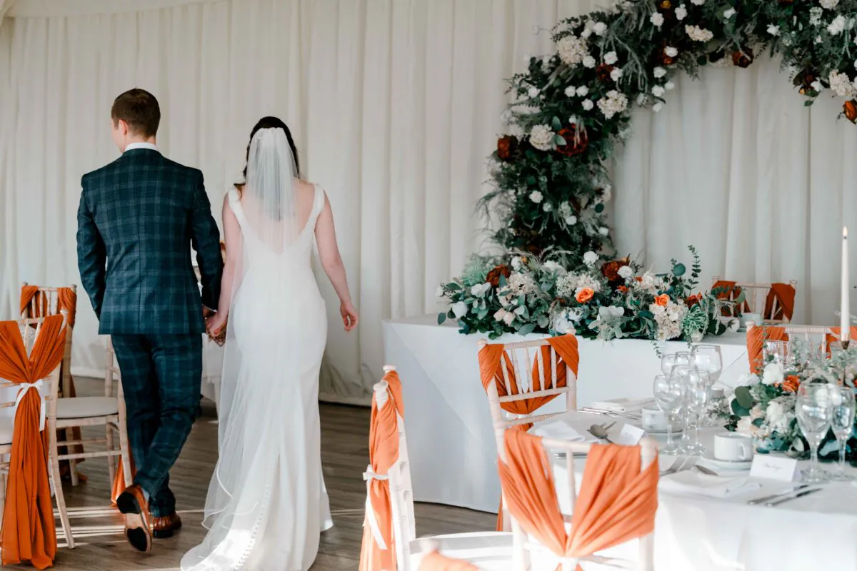 The bride and groom walk hand in hand away from the camera through a marquee reception venue decorated with burnt orange sashes and a large floral arch.