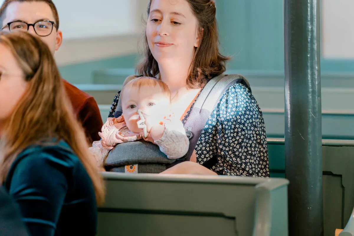 A guest sits in a church pew holding a baby in a front carrier during the wedding ceremony.