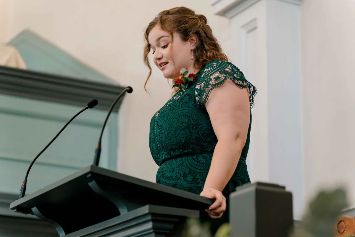 A woman in a dark green lace dress stands at a podium with two microphones, speaking during a wedding ceremony.
