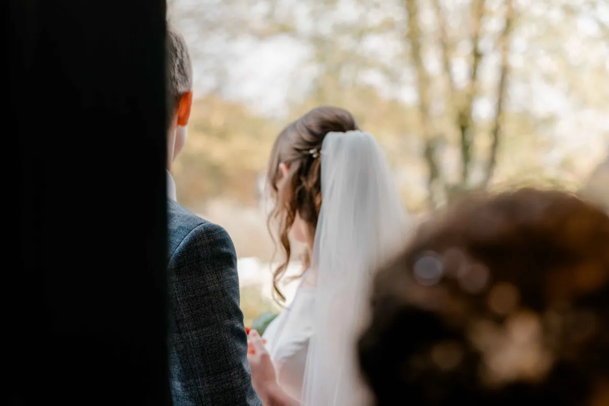 A bride wearing a chapel veil and a groom in a textured tweed suit viewed from behind through a glass window.