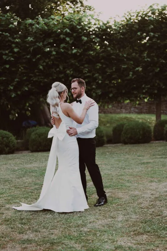 Newlyweds share a tender moment dancing together on a manicured lawn framed by lush green trees during golden hour.