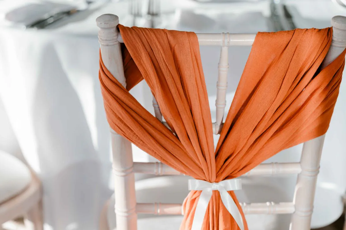 A wooden wedding chair is elegantly draped with burnt orange fabric and tied neatly at the back with a white ribbon.