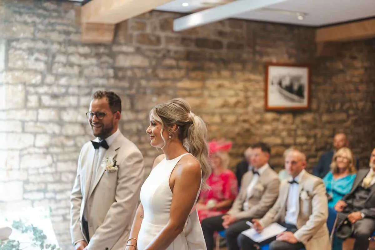 A smiling bride and groom stand together during their indoor ceremony inside a rustic stone barn, with wedding guests seated in the background.