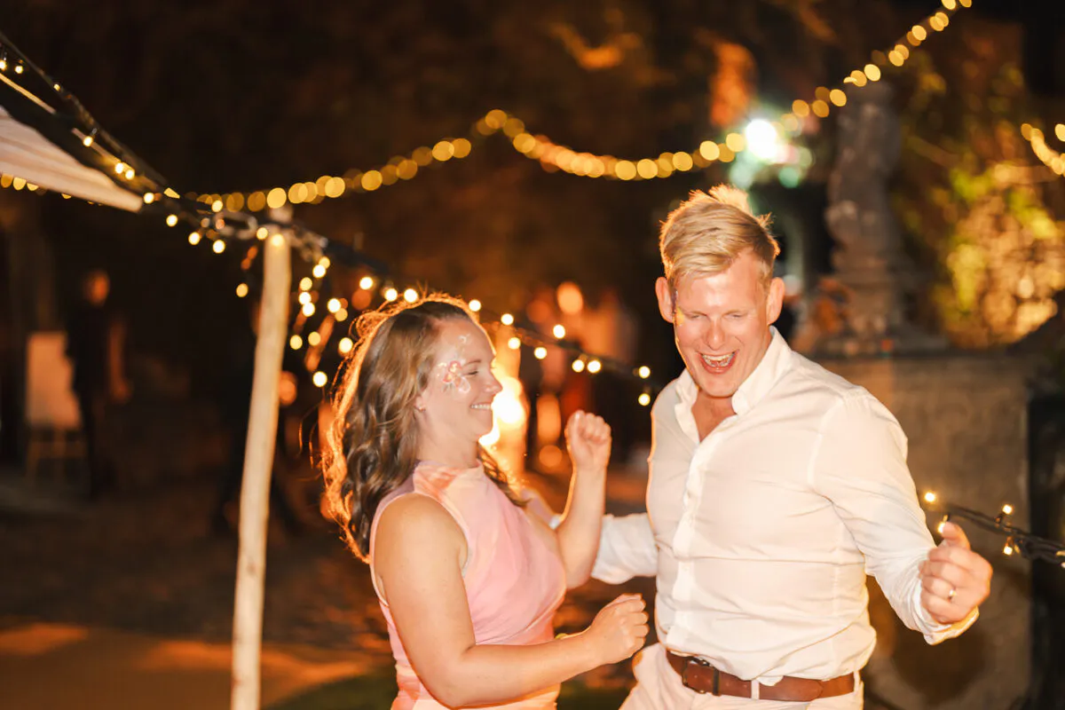 A newlywed couple laugh and dance joyfully outdoors at night beneath a glowing canopy of fairy lights.