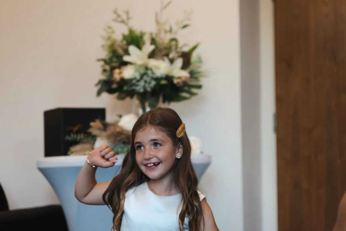 A young flower girl with long brown hair smiles playfully while raising her arm in front of a floral centrepiece arrangement.
