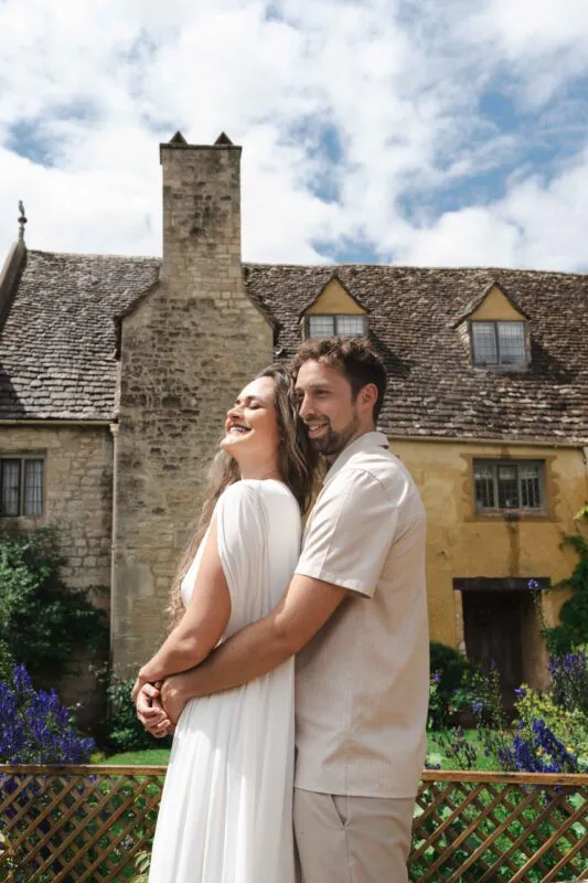 A newlywed couple embraces lovingly outdoors in front of a rustic stone manor house under a bright blue sky.