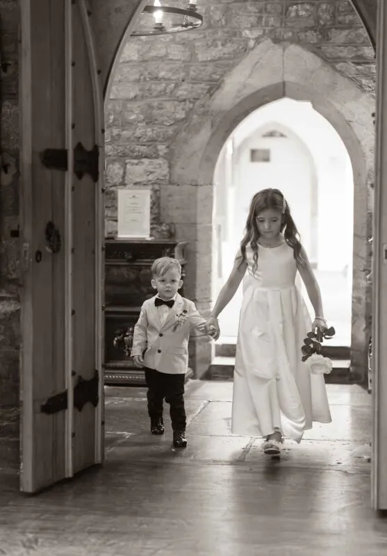 A young flower girl and page boy walk hand in hand through a stone archway in a black and white wedding photograph.