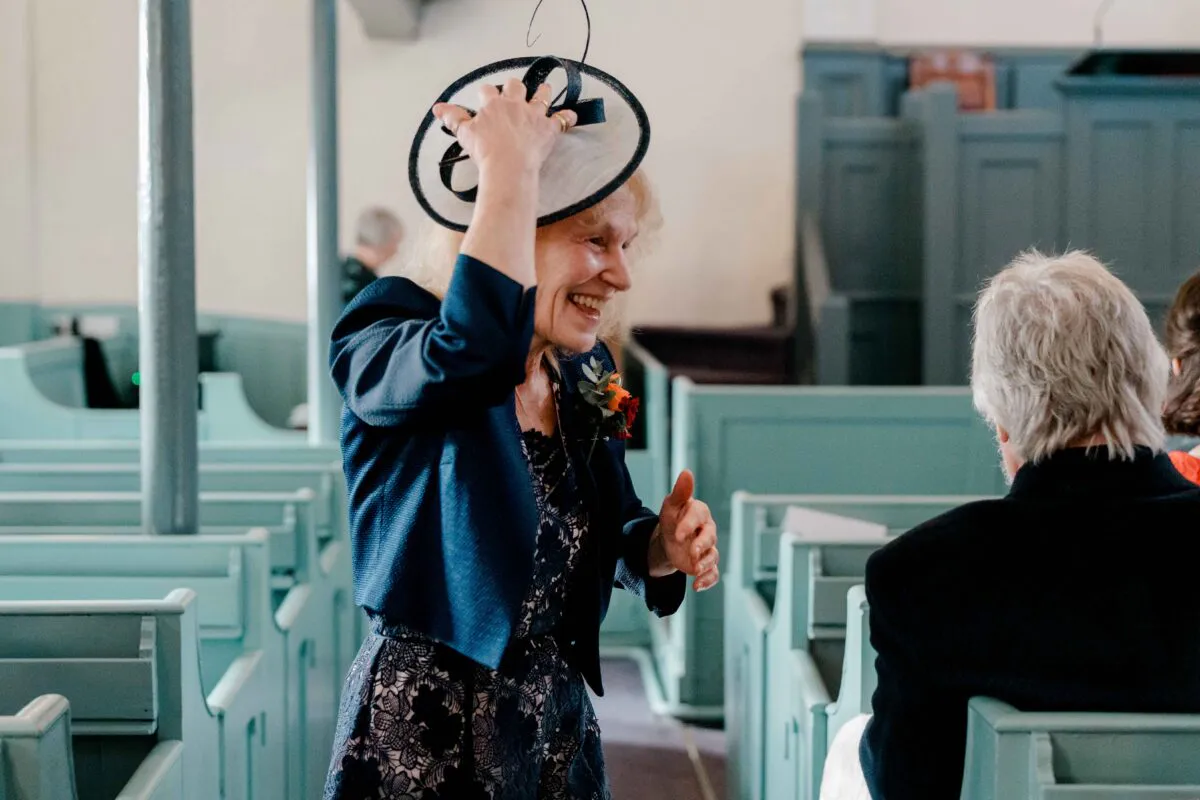 An elderly female wedding guest wearing a patterned blue dress and a black and white fascinator laughs joyfully while standing inside a church with pale blue pews.
