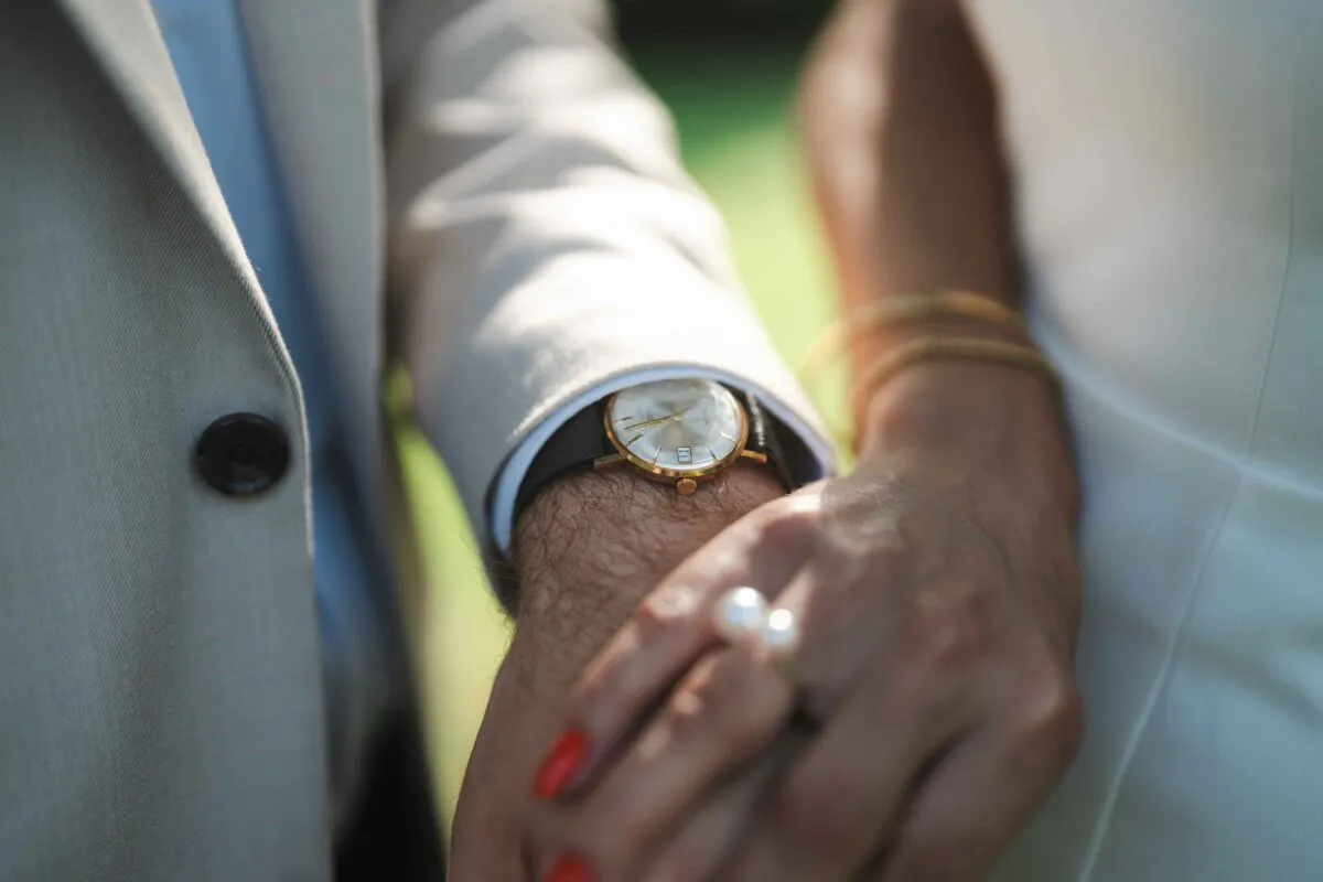 Close-up of a couple holding hands outdoors, highlighting the groom wearing a beige suit and wristwatch alongside the bride in a white outfit.
