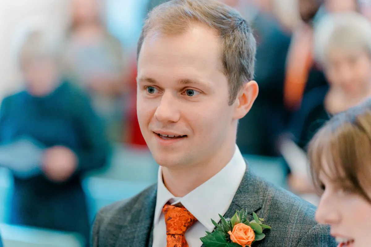 A smiling groom wearing a grey suit and orange patterned tie stands at the altar during a church wedding ceremony, with guests blurred in the background.