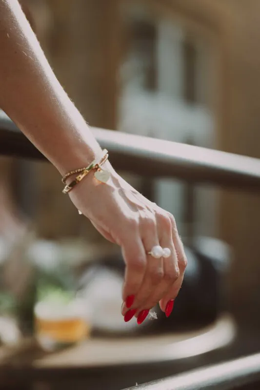 Close-up shot of a bride's hand adorned with delicate gold bracelets and a pearl statement ring, featuring vibrant red nails.