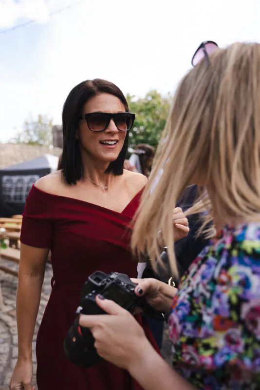 A guest wearing a burgundy off-the-shoulder dress and sunglasses smiles while chatting outdoors, viewed past another guest holding a camera.