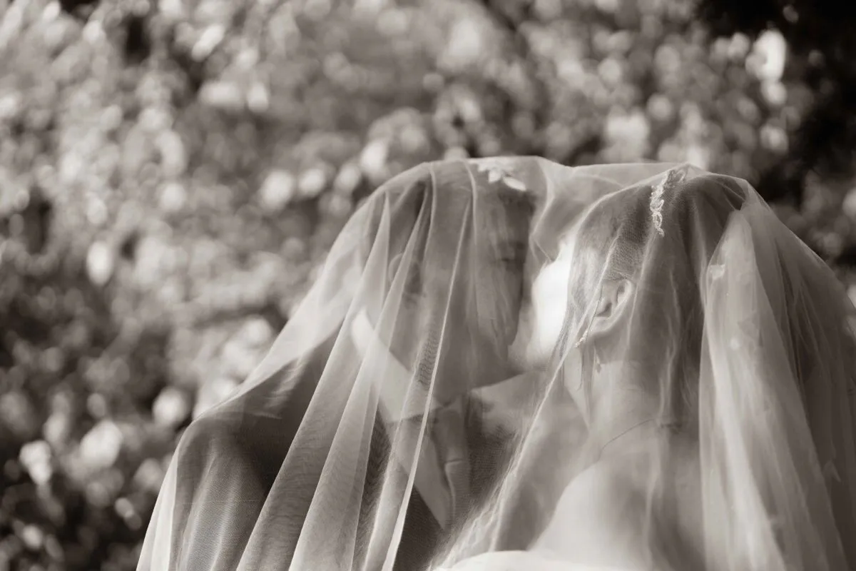 An intimate black and white portrait captures a bride and groom sharing a tender kiss beneath a shared veil against a backdrop of soft foliage.