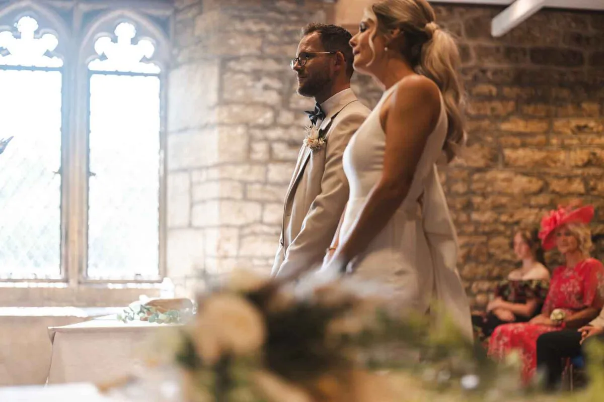 A bride and groom stand side by side during their indoor ceremony inside a stone-walled venue illuminated by bright window light.
