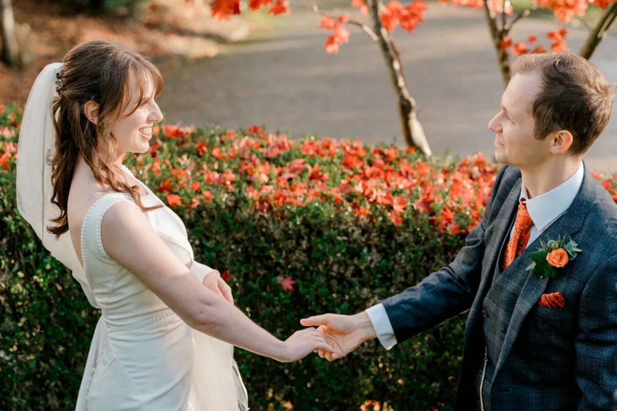 A newlywed couple holds hands and smiles at each other outdoors against a vibrant backdrop of autumn foliage.