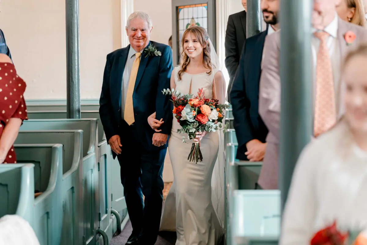 A smiling bride walks down the church aisle holding her father's arm while carrying a colourful autumnal bouquet.
