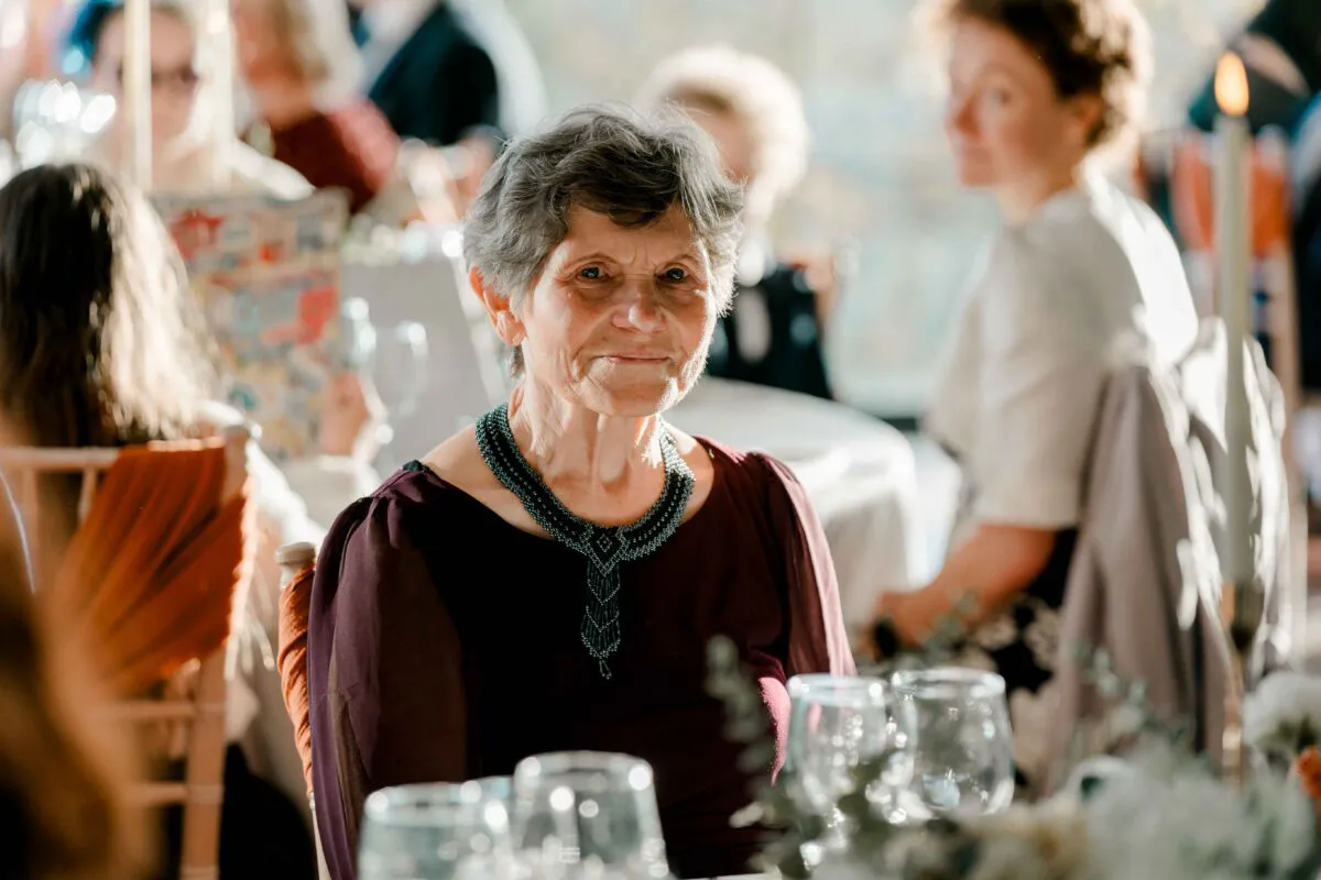 An elderly female wedding guest smiles warmly while seated at a reception table amidst glasses and floral decor.