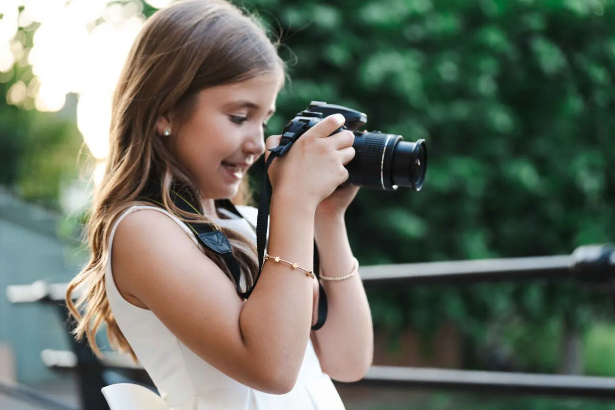 A young flower girl smiling while holding a DSLR camera outdoors with a lush green backdrop.