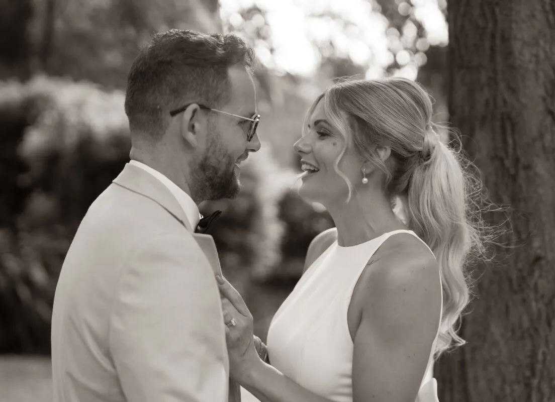 A newlywed couple share a joyful, laughing moment face-to-face outdoors against a softly blurred woodland backdrop.