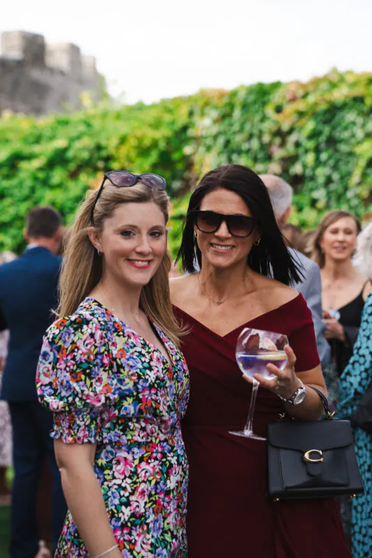 Two wedding guests smile together outdoors during the drinks reception, with one wearing a vibrant floral dress and sunglasses on her head.