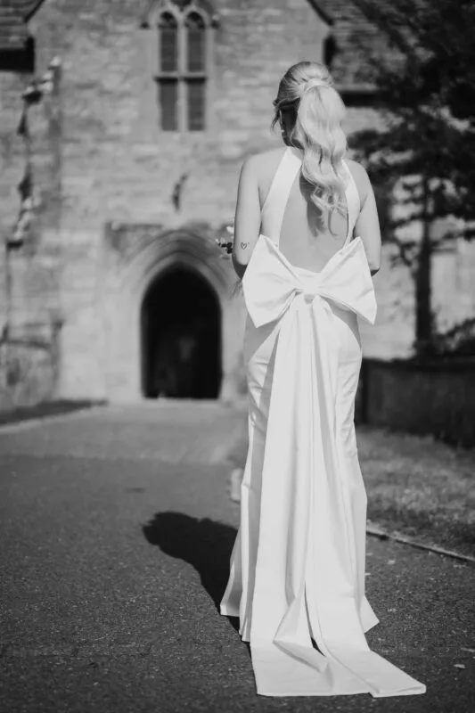 A stunning bride viewed from behind stands on a pathway outside a historic stone church, wearing a chic halter-neck gown featuring a dramatic oversized bow and long train.