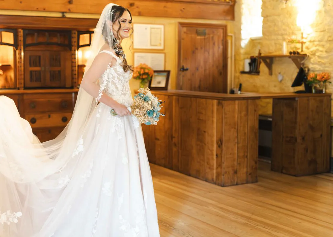 A smiling bride wearing an a-line lace wedding dress and veil holds a dried flower bouquet inside a rustic barn venue with wooden paneling.