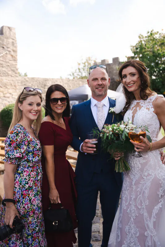 The bride and groom pose smiling outdoors at a castle venue alongside wedding guests holding drinks during a sunny drinks reception.