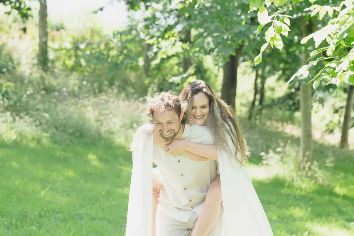 A joyful newlywed couple share a playful piggyback ride amidst a sunlit British woodland on their wedding day.