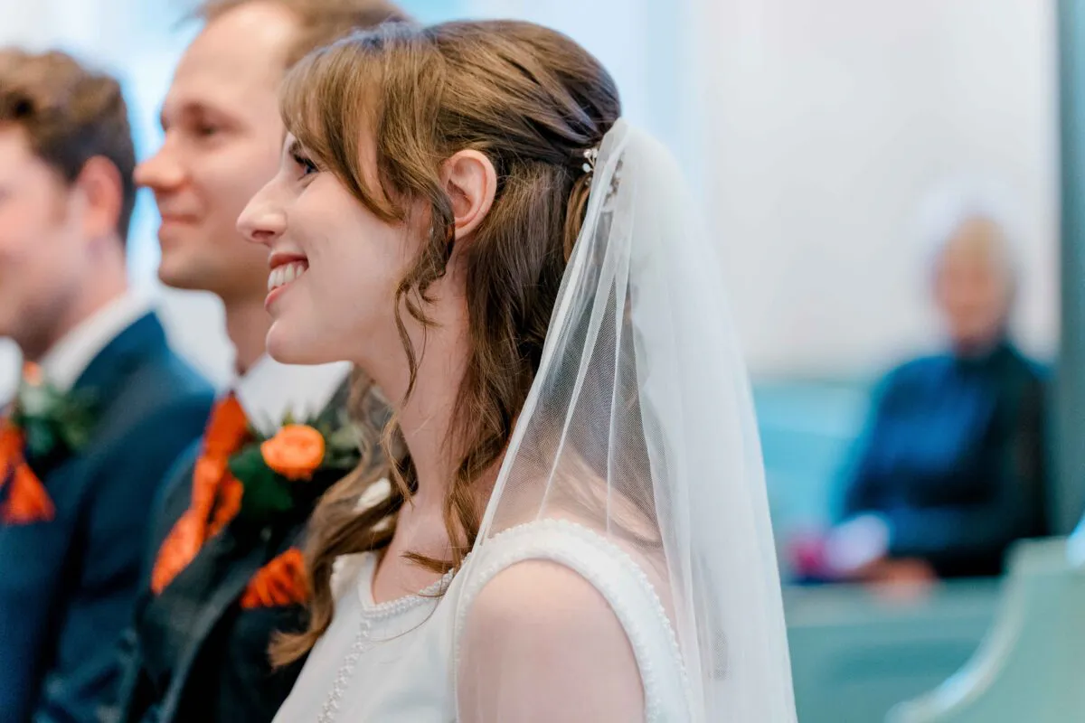 A smiling bride wearing a half-up hairstyle and a veil stands alongside her groom during the wedding ceremony.