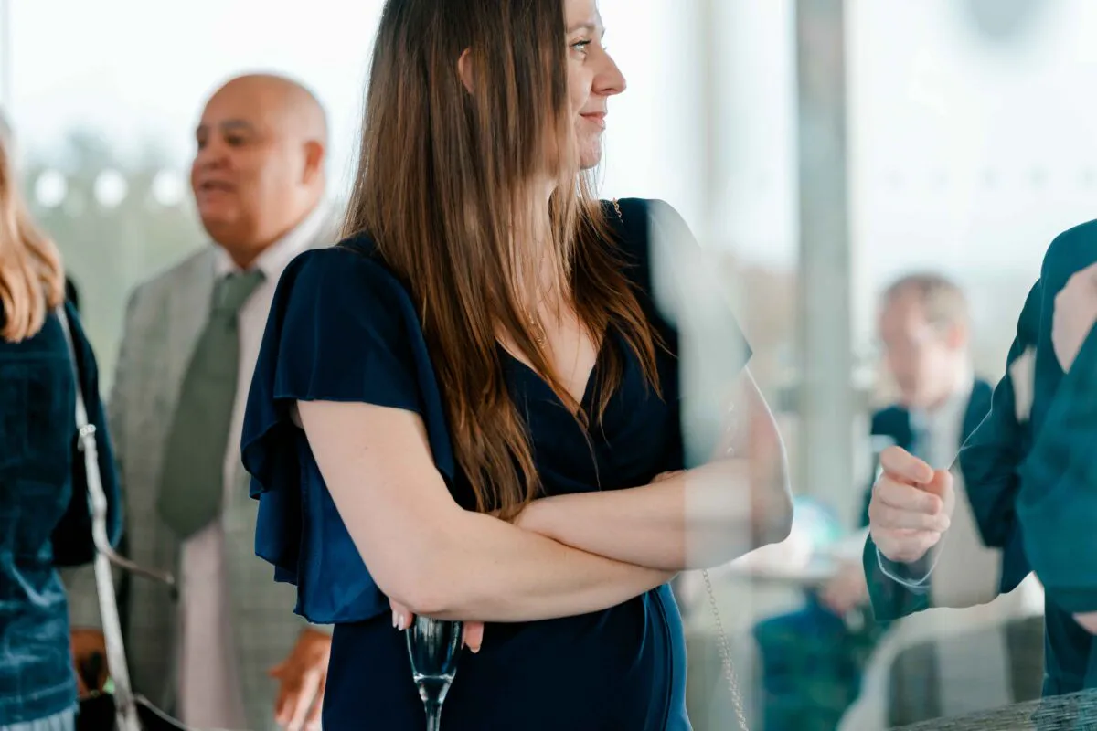 A wedding guest in a dark blue dress stands with her arms crossed while mingling during a drinks reception.